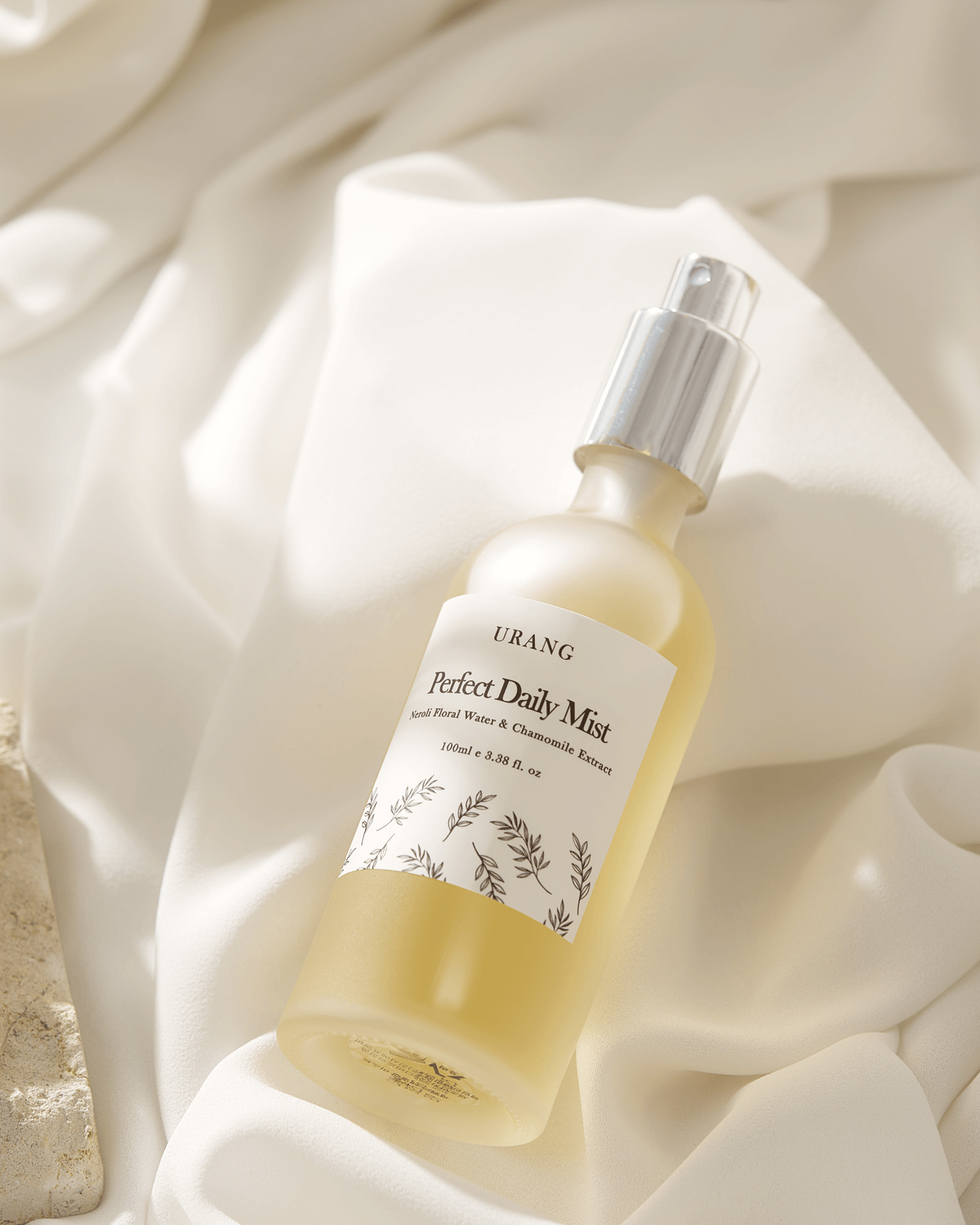 Close-up of the Urang Perfect Daily Mist frosted glass bottle, with a silver cap and a yellow-tinted liquid inside, resting diagonally on soft, cream-colored, draped fabric.