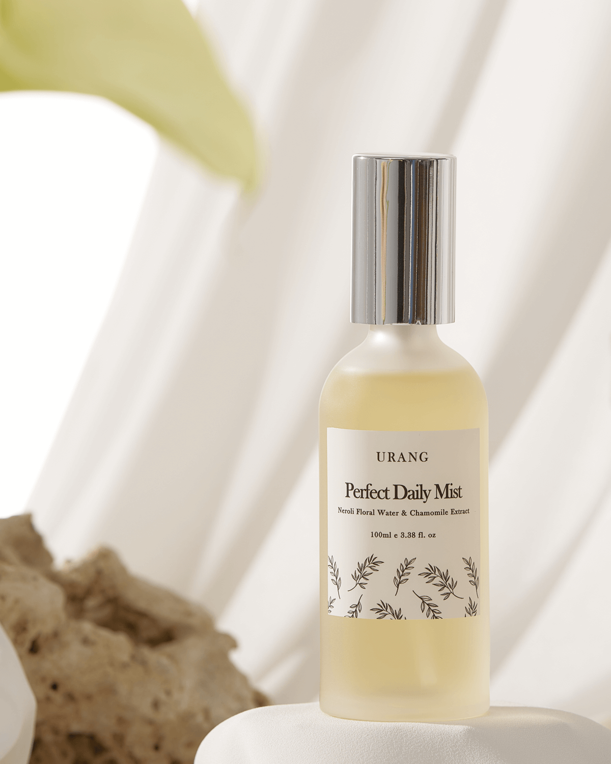 The frosted glass Urang Perfect Daily Mist bottle with a silver cap, standing on a white, textured pedestal, surrounded by soft white fabric and receiving bright, angled studio lighting.