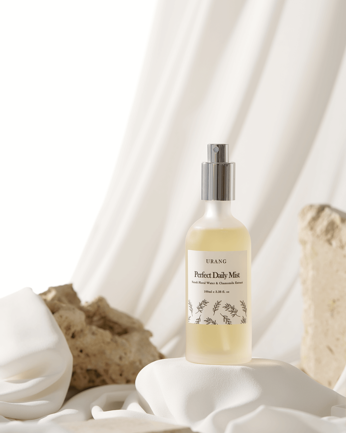 The Urang Perfect Daily Mist frosted glass bottle, containing a pale yellow liquid, displayed on a textured white pedestal and surrounded by soft white fabric and natural stones.