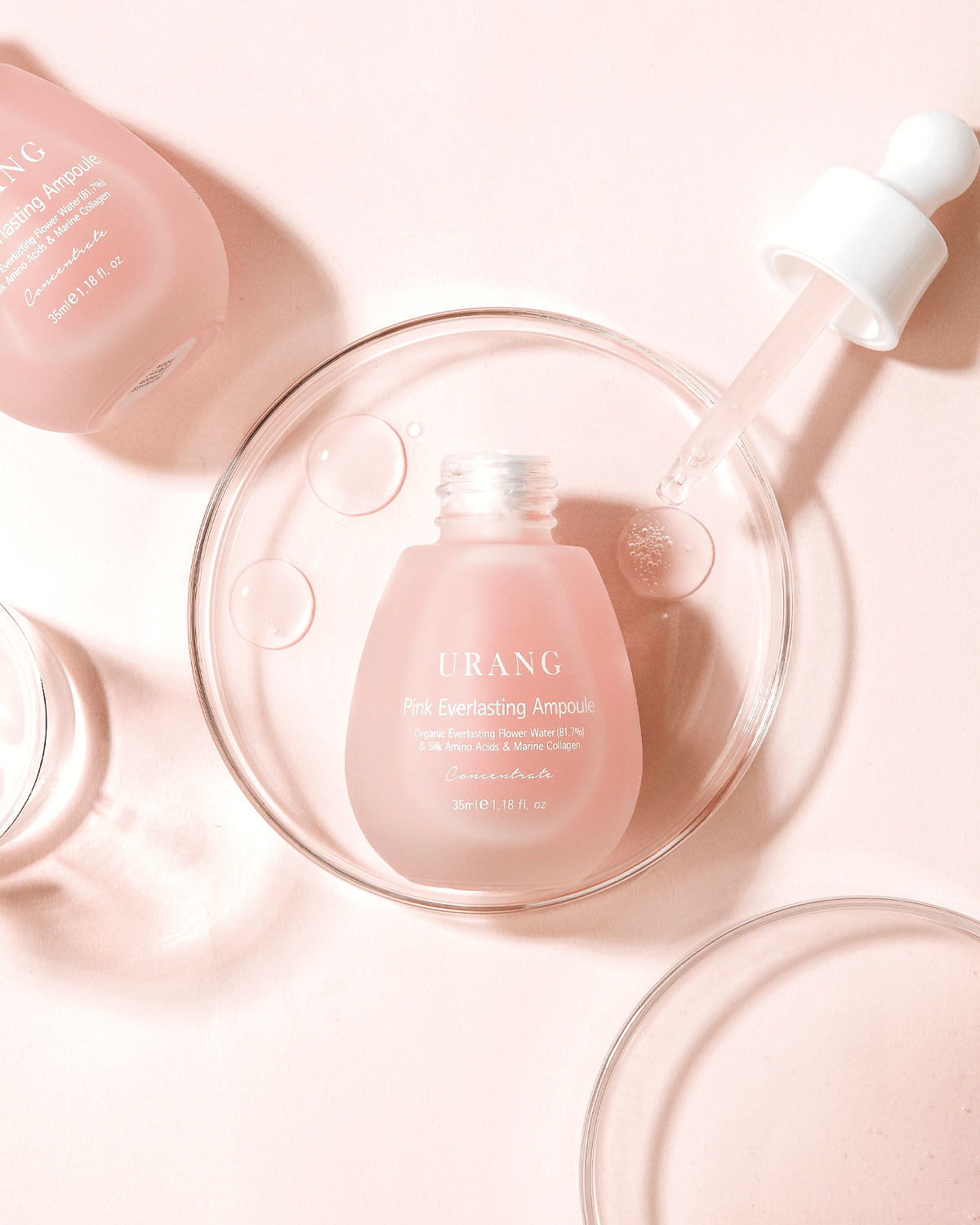 Top-down view of the Urang Pink Everlasting Ampoule, a frosted pink glass dropper bottle sitting in a clear, circular laboratory dish on a white background, with another bottle blurred in the top-left corner.