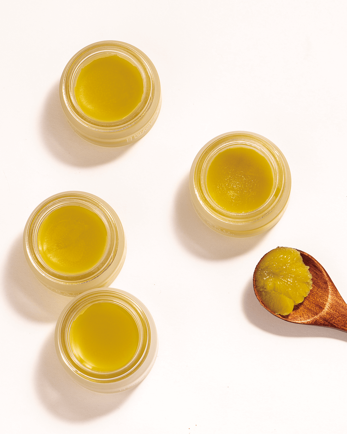 Overhead shot of four open, clear glass jars of Urang S.O.S. Multi Balm with Tamanu, showcasing the solid, pale yellow-green balm, next to a wooden spoon holding an additional scoop of the product.