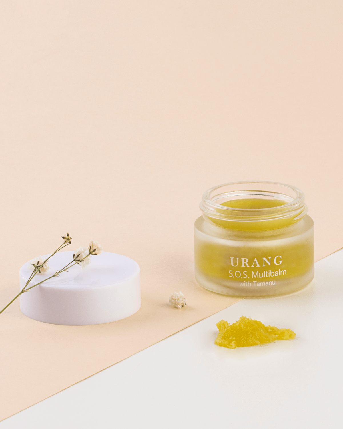 The Urang S.O.S. Multi Balm with Tamanu in a clear glass jar, with a detached white cap and a scoop of the solid yellow-green balm resting on a white surface, complemented by a delicate dried flower stem.