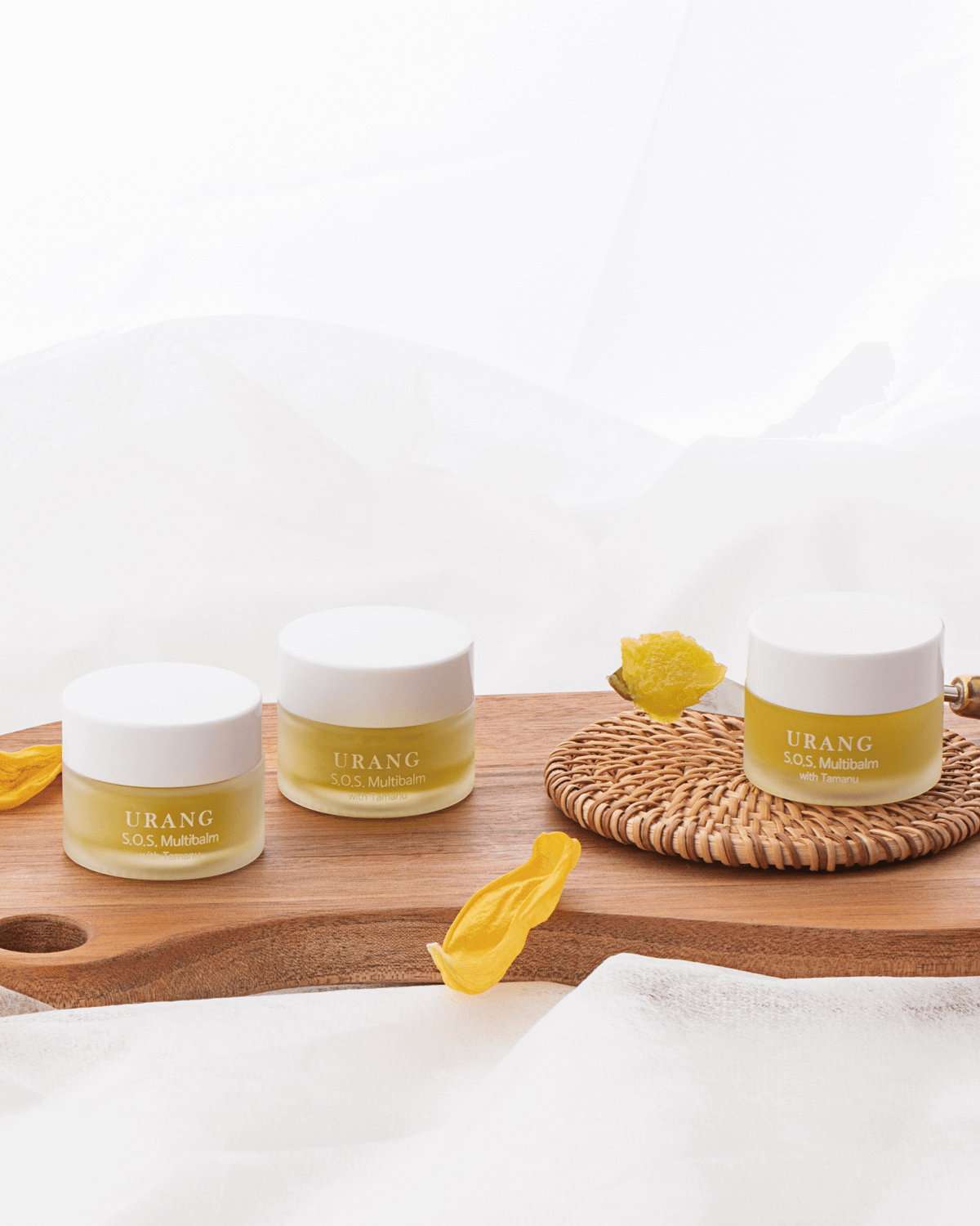 Three jars of the Urang S.O.S. Multi Balm with Tamanu, showcasing the pale yellow-green balm, arranged on a round, woven wicker mat, with dried yellow flower petals scattered on a wooden cutting board.