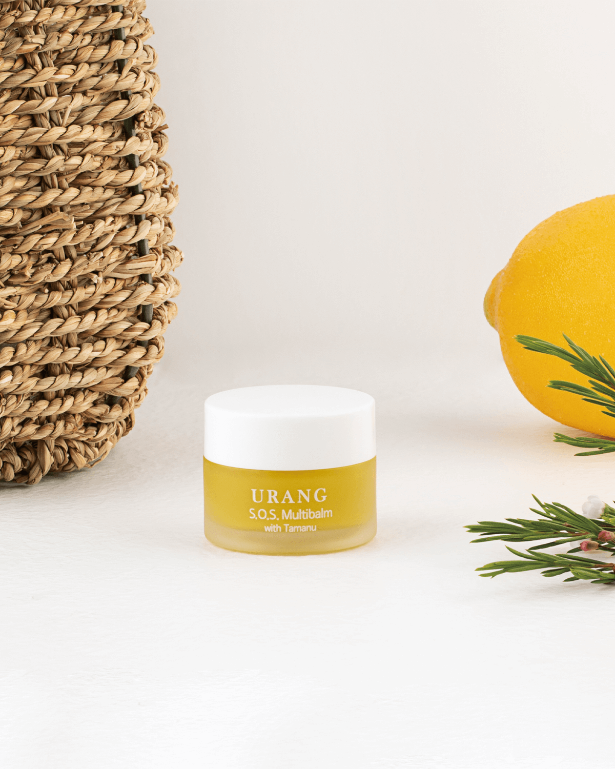 The small, frosted glass jar of Urang S.O.S. Multi Balm with Tamanu, showcasing the pale yellow balm, is placed on a white surface next to a woven basket, a bright yellow lemon, and a sprig of greens.