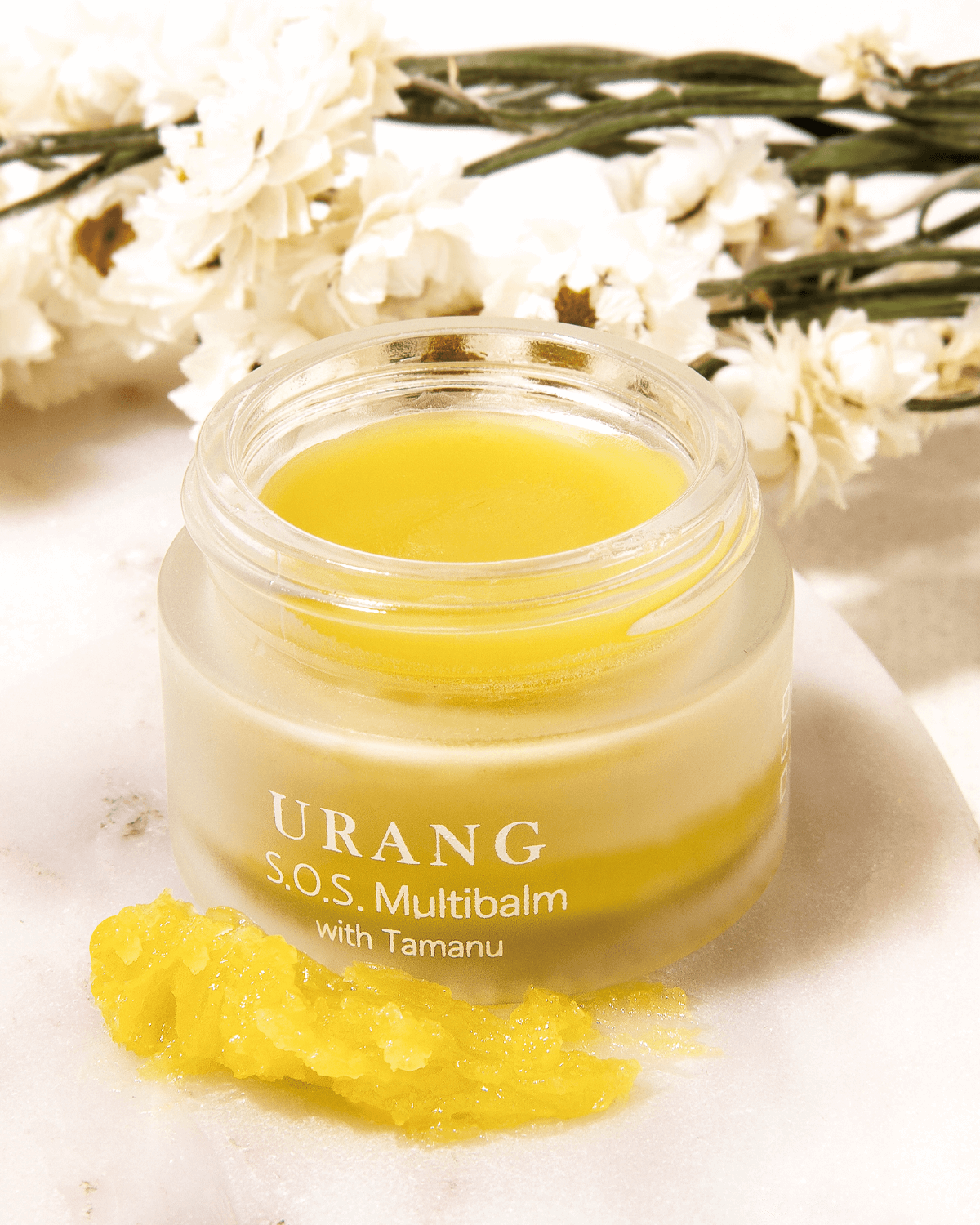 Close-up of the Urang S.O.S. Multi Balm with Tamanu in a wide glass jar, showing the smooth, pale yellow-green solid balm, with a scoop of the balm resting beside the jar and white dried flowers in the blurred background.
