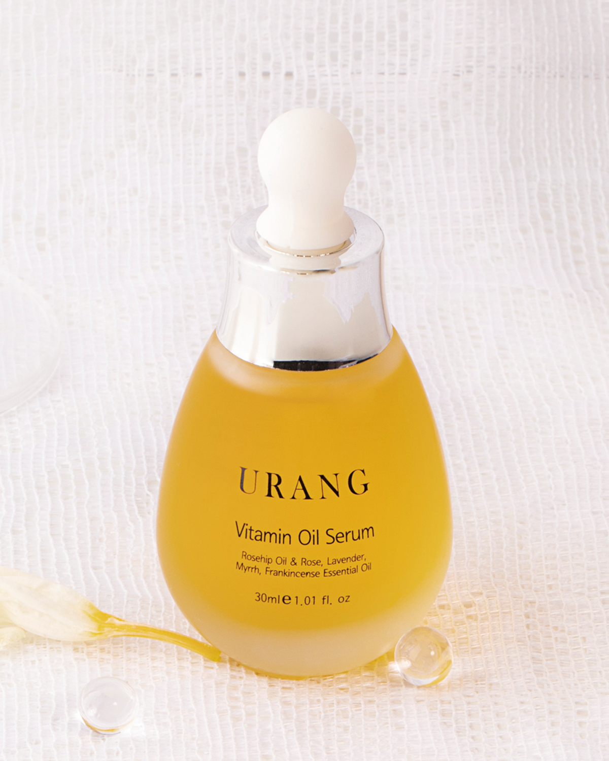 The Urang Vitamin Oil Serum in a frosted glass dropper bottle, featuring the bright yellow-orange oil, is displayed on a textured white fabric with a couple of tiny glass balls.