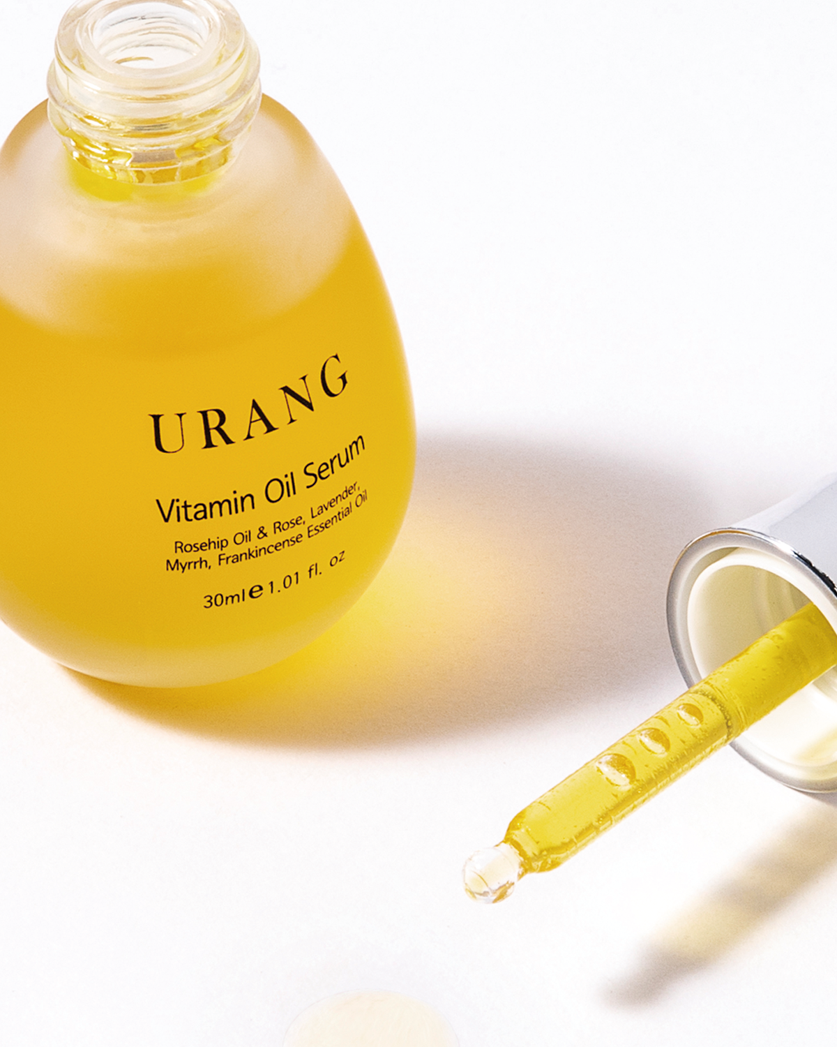 Close-up of the Urang Vitamin Oil Serum in its frosted glass dropper bottle, featuring the vibrant yellow-orange oil. The glass pipette is drawn out, with a drop of oil visible at the tip, resting on the white surface.