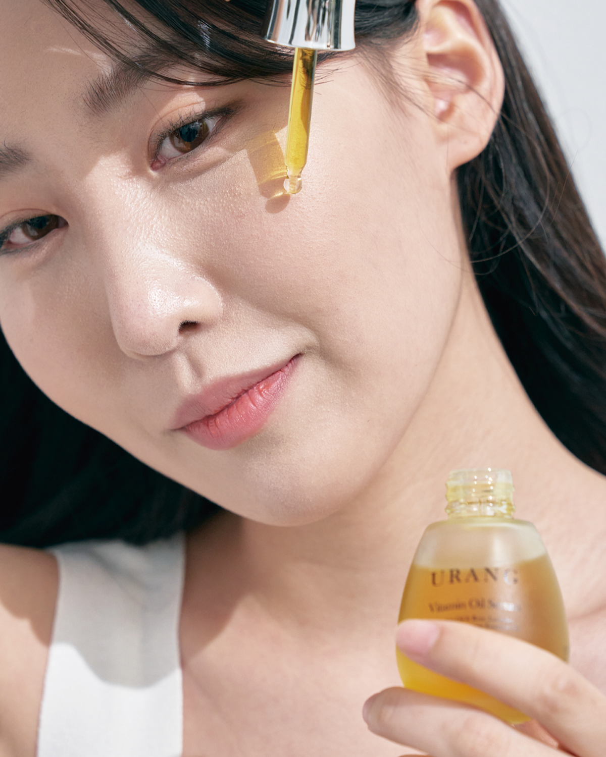 Close-up portrait of a woman holding the frosted glass Urang Vitamin Oil Serum bottle, with a drop of the vibrant yellow-orange oil visibly suspended from the glass dropper tip near her cheek, ready for application.
