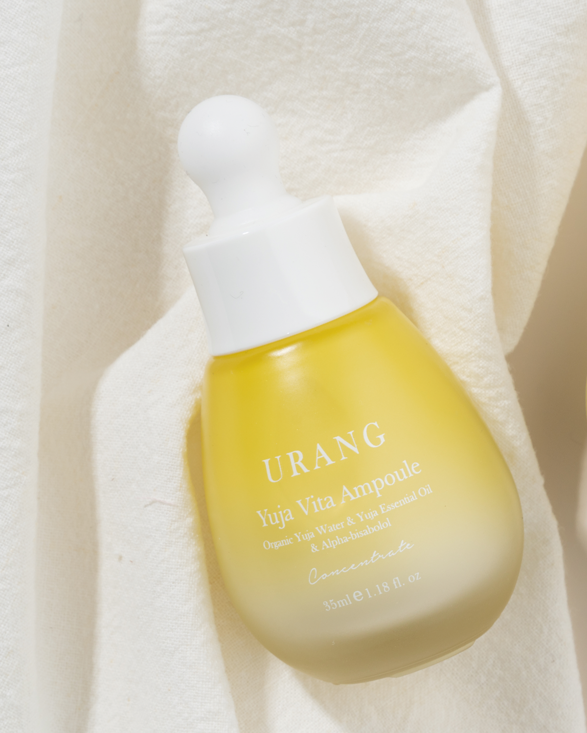 Close-up, angled view of the Urang Yuja Vita Ampoule, a milky, pale yellow serum in a frosted glass dropper bottle, standing on a white linen or textured fabric background.