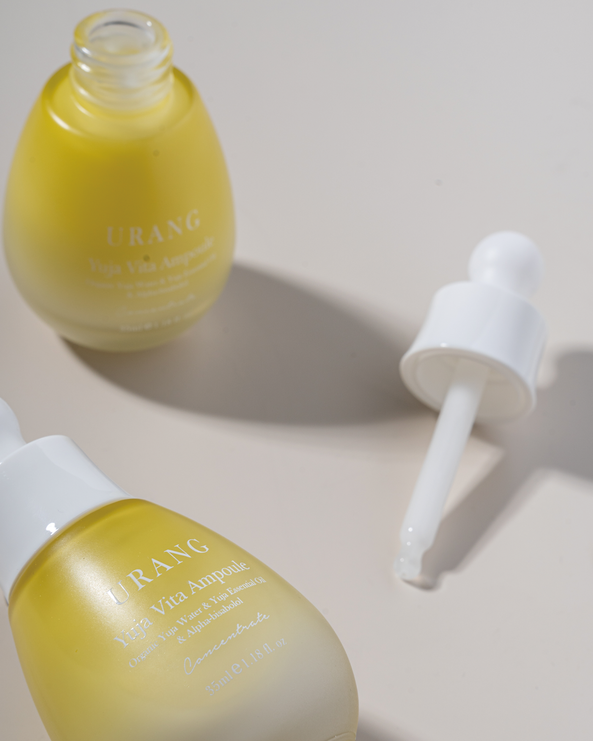 Close-up of two Urang Yuja Vita Ampoule bottles: one upright and one tilted, showcasing the milky, pale yellow serum inside and the white dropper resting on the light background.