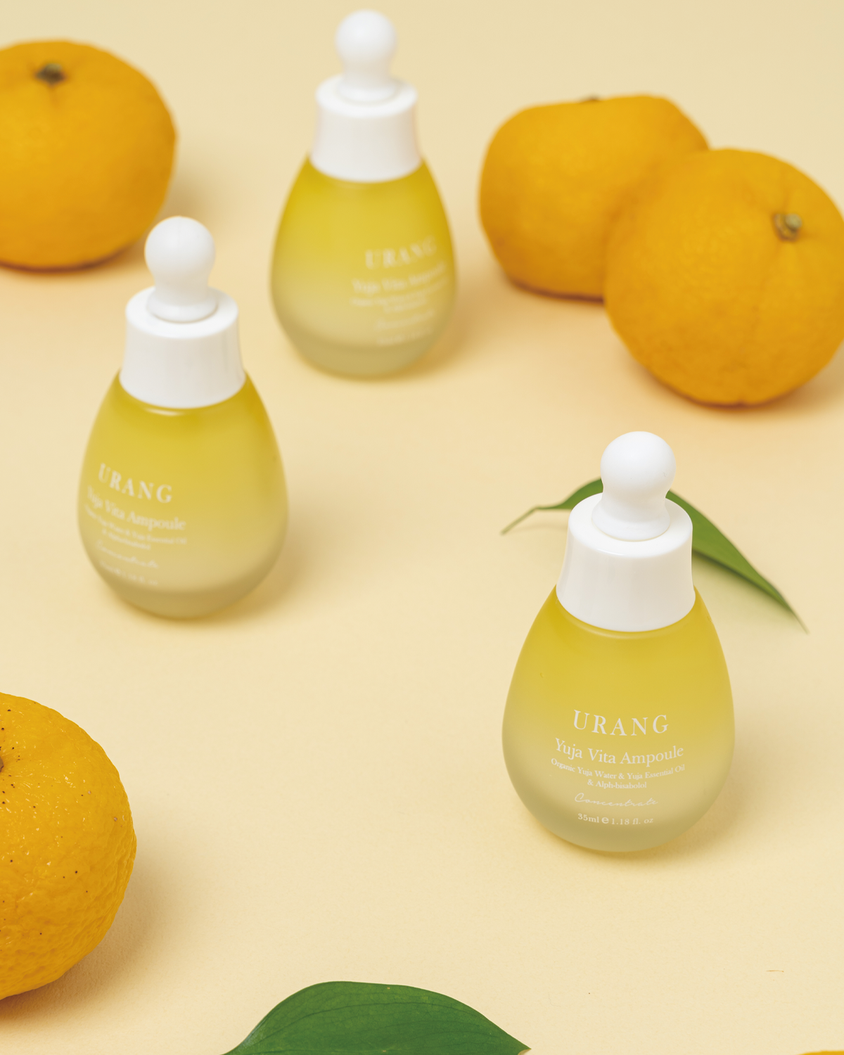 Three frosted glass bottles of Urang Yuja Vita Ampoule, showing the milky yellow serum inside, arranged on a light surface with three whole Yuja fruits and a green leaf as props.