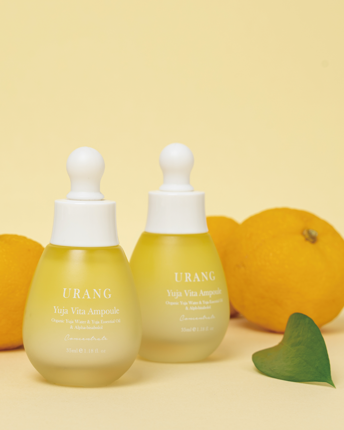 Two frosted glass bottles of Urang Yuja Vita Ampoule, showing the milky yellow serum inside, arranged with two whole Yuja fruits and a green leaf on a light yellow background.