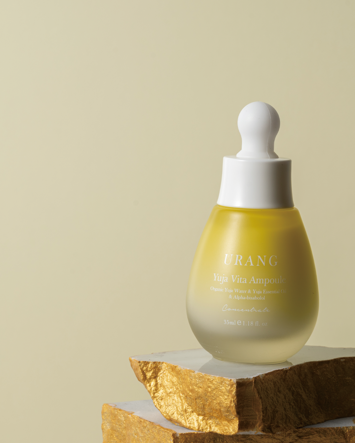 The Urang Yuja Vita Ampoule, a milky yellow serum in a frosted glass dropper bottle, standing on a stack of rough, gold-painted rocks against a pale yellow background.