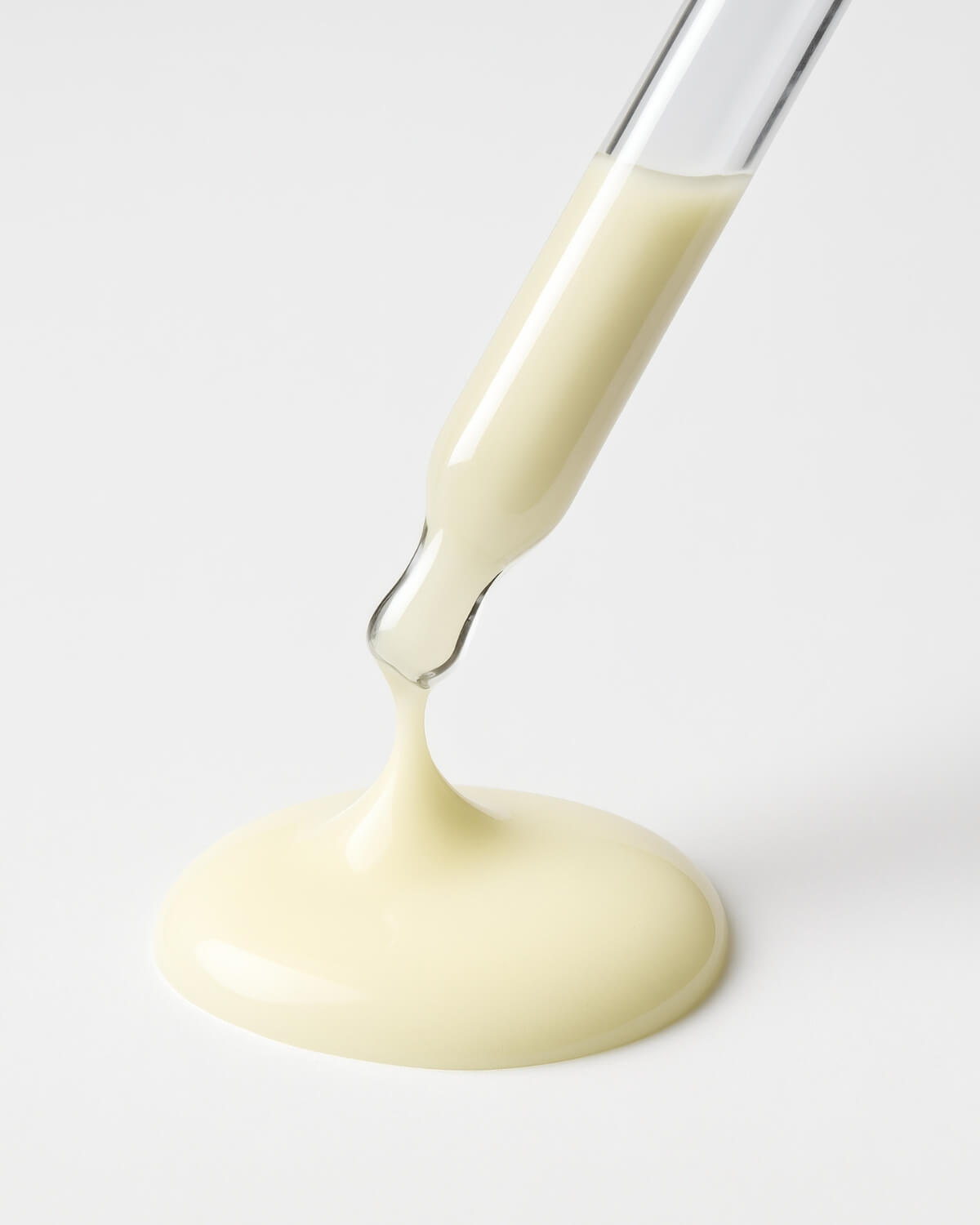 A close-up shot of a glass dropper from which a thick stream of pale yellow serum is pouring and pooling into a large, glossy circle on a white surface.