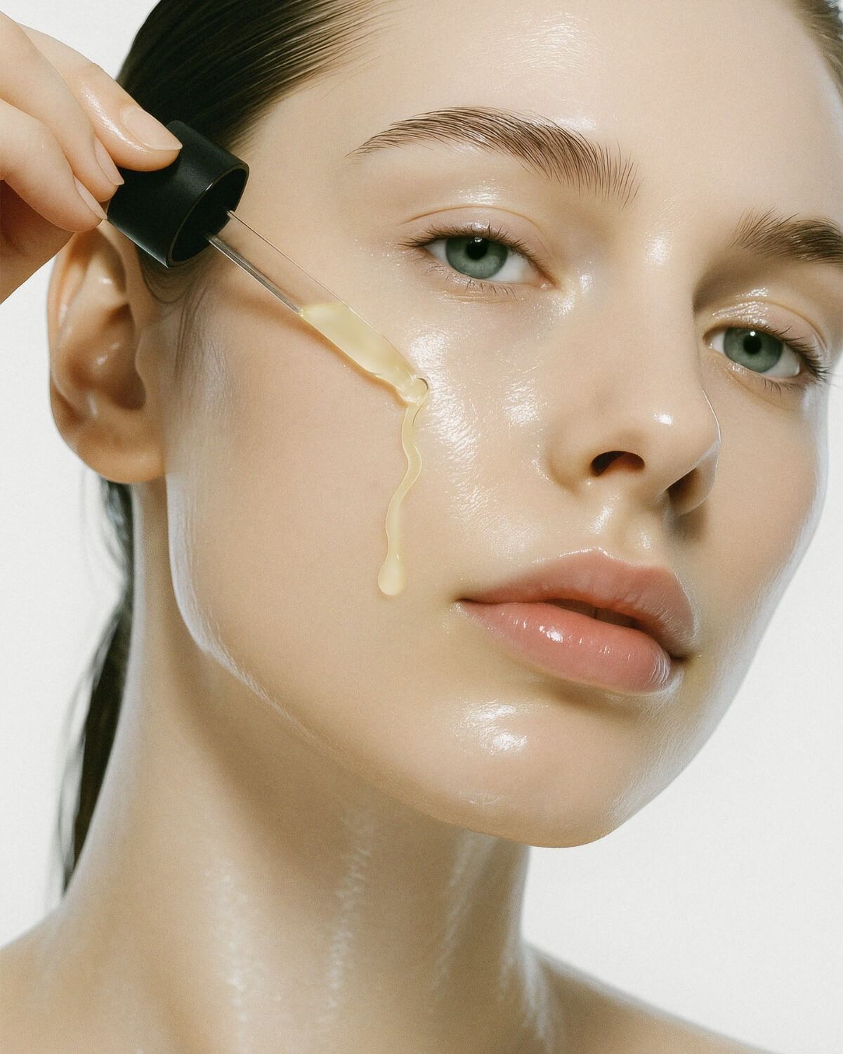 A close-up of a woman's face with highly moisturized, dewy skin. A hand holds a dropper, releasing a stream of pale yellow serum that runs in a wavy line down her cheek.