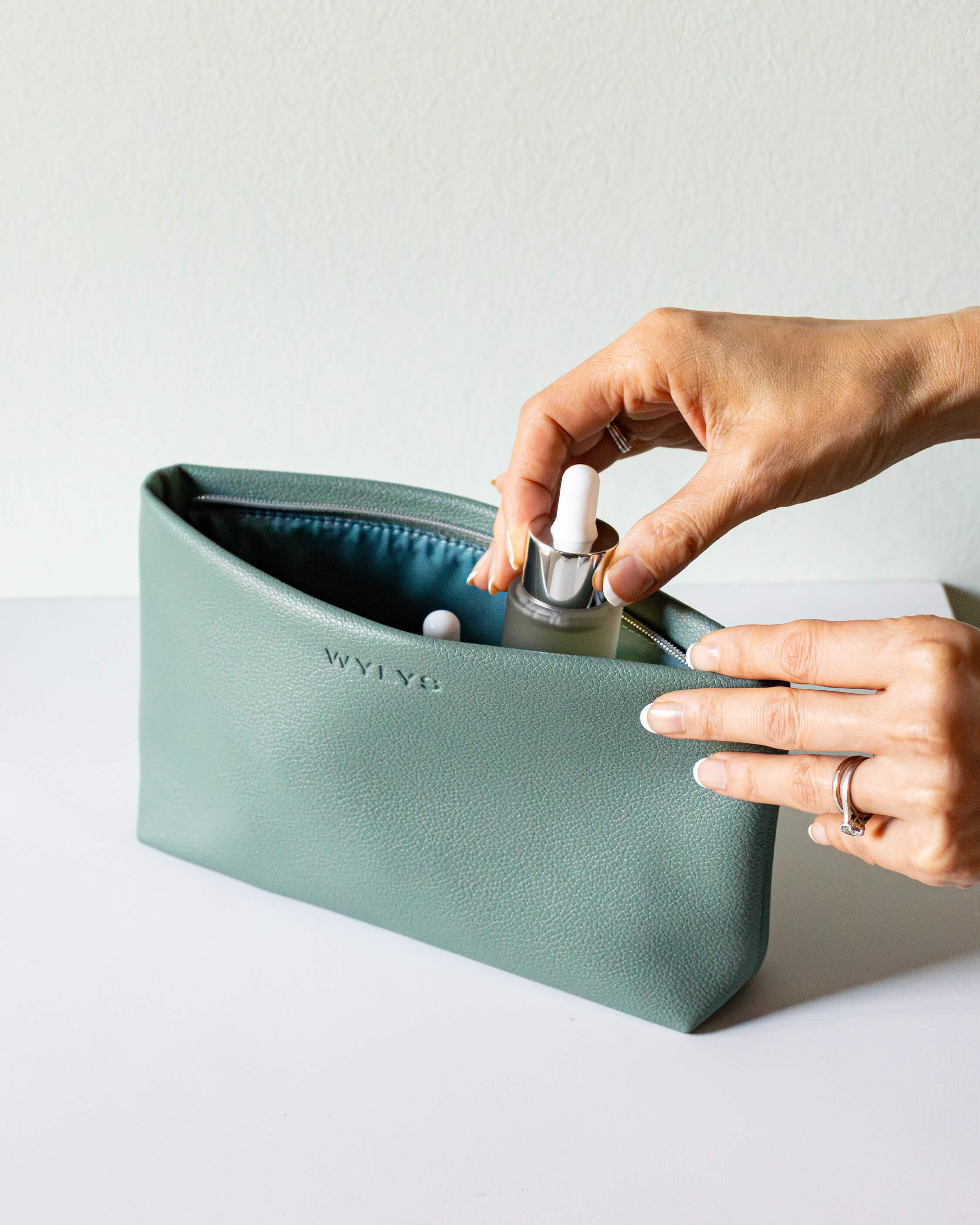 WYLYS Sage Verte Makeup Bag Soft Vegan Leather Water-resistant Hands placing skincare into sage makeup bag. Hold skincare makeup. 9.25” x 5” x 2”