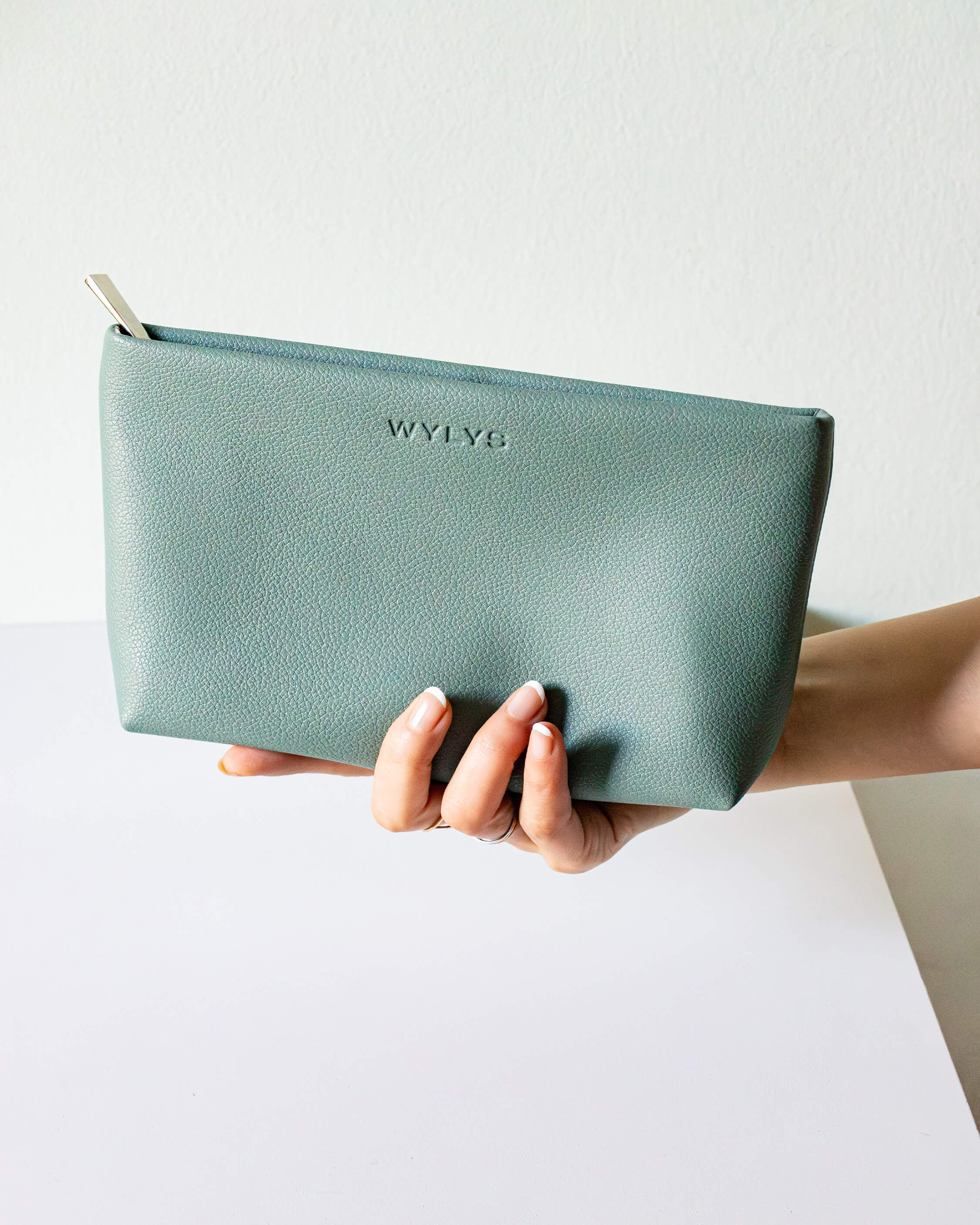 WYLYS Sage Verte Makeup Bag Soft Vegan Leather Water-resistant Semi-closeup Metal pull Debossed WYLYS logo on bag. Hand holding bag.