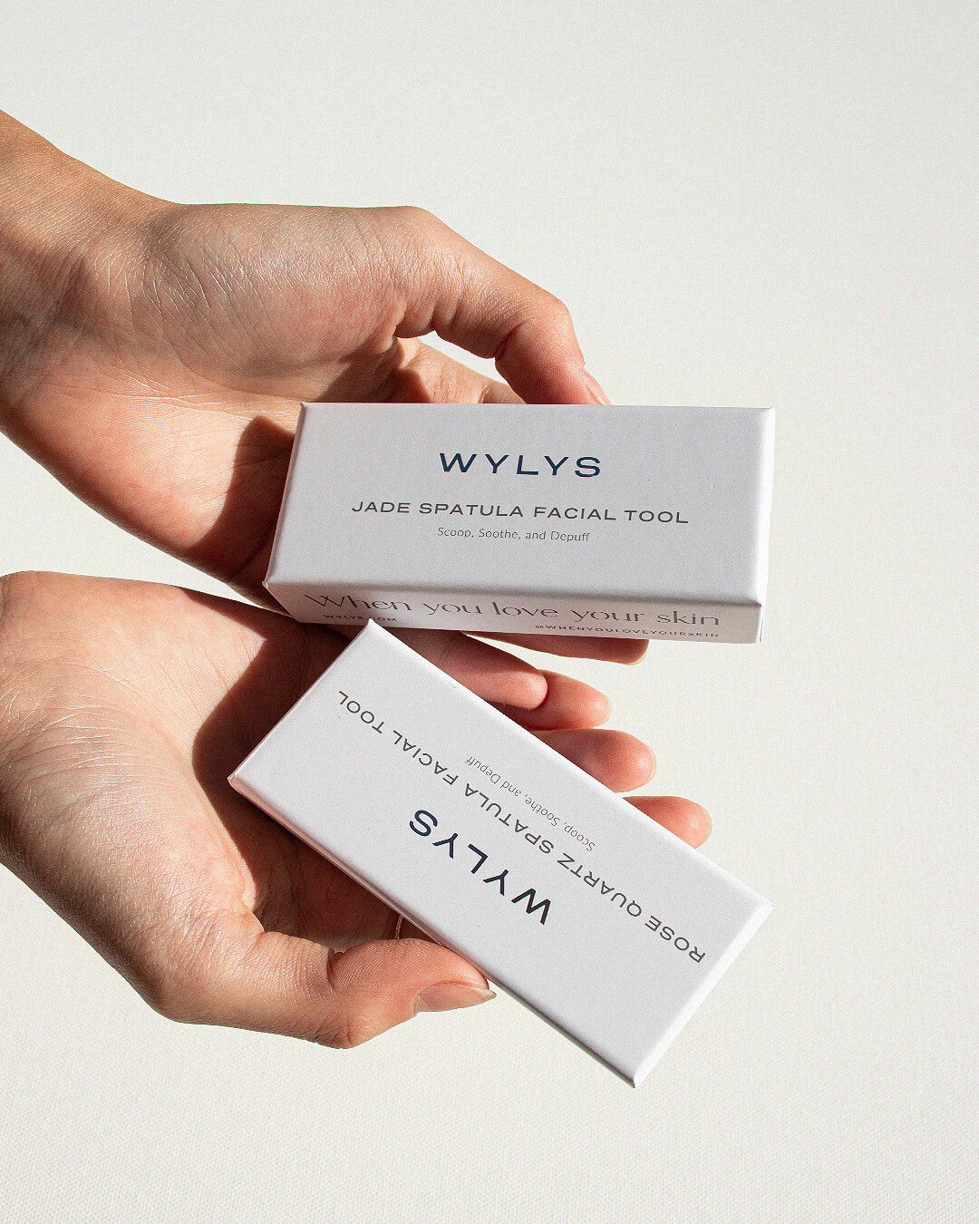 OHLOLLY Korean Skincare Online Shop WYLYS Rose Quartz and Jade Spatula Facial Tool Facial Tool to Scoop, Soothe, and Depuff 100% Natural Stone, Massage, Ergonomic Skincare Spatula to use with Creams & Balms. Gift-friendly. Hands holding two closed boxes.