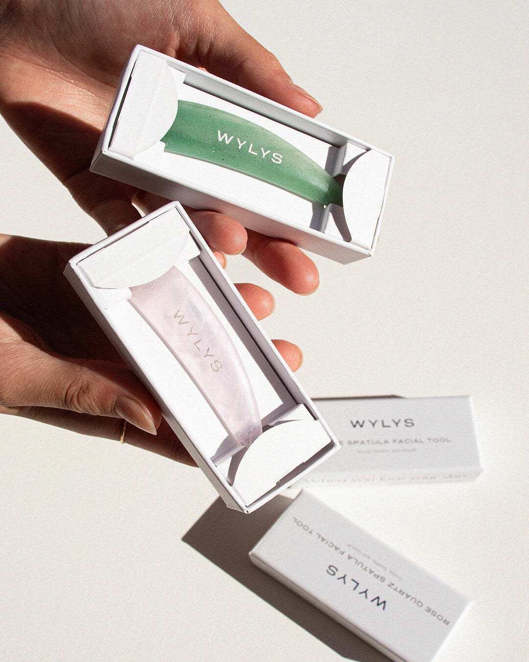 OHLOLLY Korean Skincare Online Shop WYLYS Rose Quartz and Jade Spatula Facial Tool Facial Tool to Scoop, Soothe, and Depuff 100% Natural Stone, Massage, Ergonomic Skincare Spatula to use with Creams & Balms. Gift-friendly. Hands holding open boxes showing products inside.