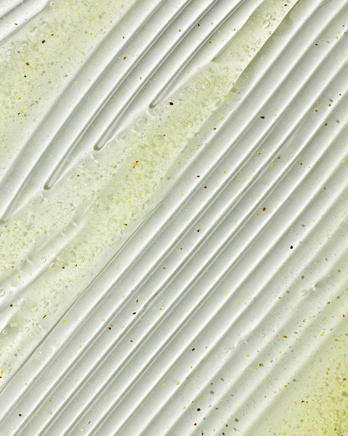 Abstract close-up of the yellowish gel cleanser texture, showing diagonal ridges, air bubbles, and small dark exfoliating specks.