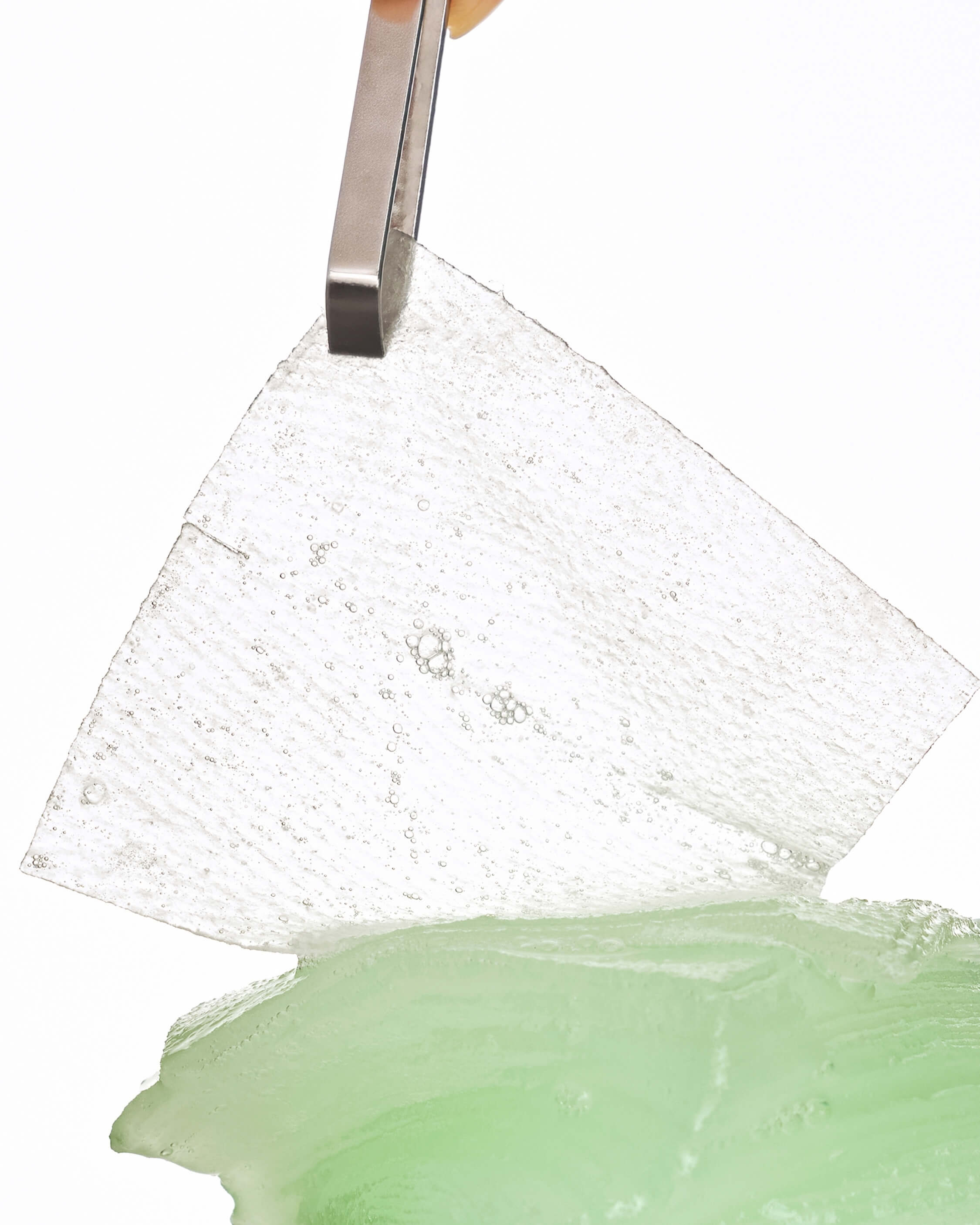 Close-up of a pair of metal tweezers lifting a clear, textured gel pad with bubbles from a pile of green gel material.