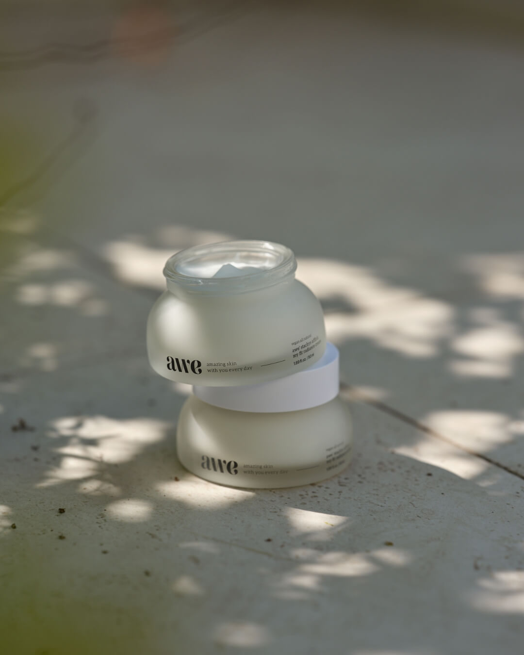 Open jar of Awe daily radiance cream stacked on top of a closed jar in natural leaf shadows.