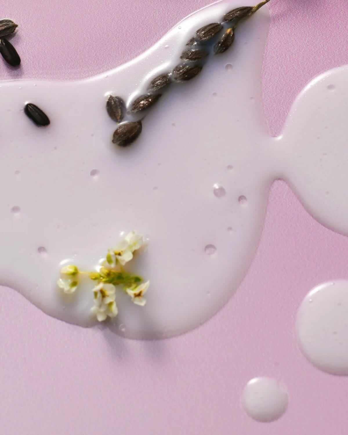 A close-up shot of milky white serum droplets on a light purple surface, accented with black rice grains and tiny white flowers.