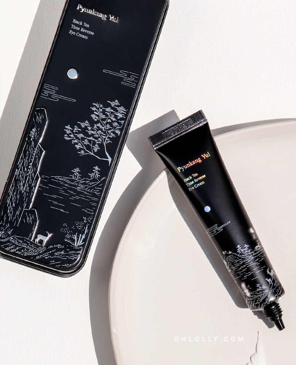 Angled studio shot of the black Pyunkang Yul Black Tea Time Reverse Eye Cream tube lying on a white dish, positioned next to its black box, which features an elegant white landscape illustration. The image highlights the product's premium, anti-aging focus with black tea.