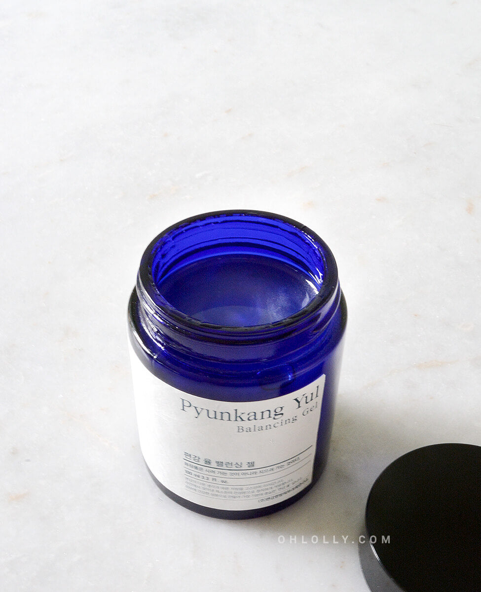 Overhead close-up of the open, dark blue Pyunkang Yul Balancing Gel jar on a white marble surface. The clear, honey-like gel is visible inside the jar, emphasizing its thick, concentrated texture and minimalist packaging.