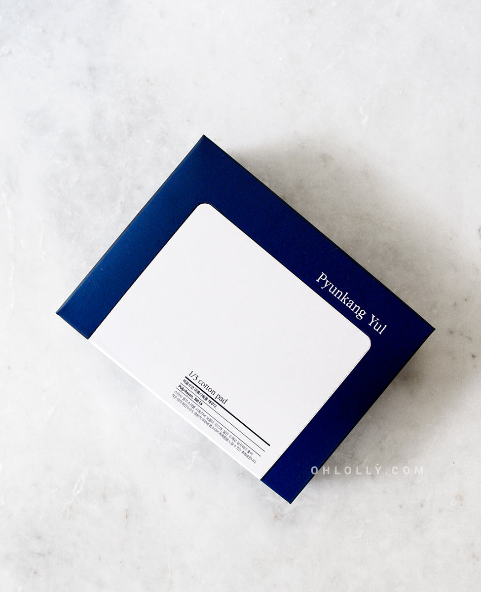 Usage shot of a hand holding up a single, thin, white Pyunkang Yul 1/3 Cotton Pad against a dark background, showing its smooth texture and highlighting its thinness for use with less toner or essence.