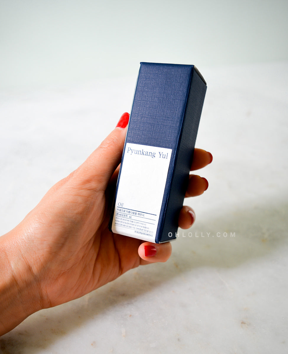 A hand with painted nails holds the dark blue, rectangular packaging box for the Pyunkang Yul Oil. The white label on the box, featuring the product name, is clearly visible.