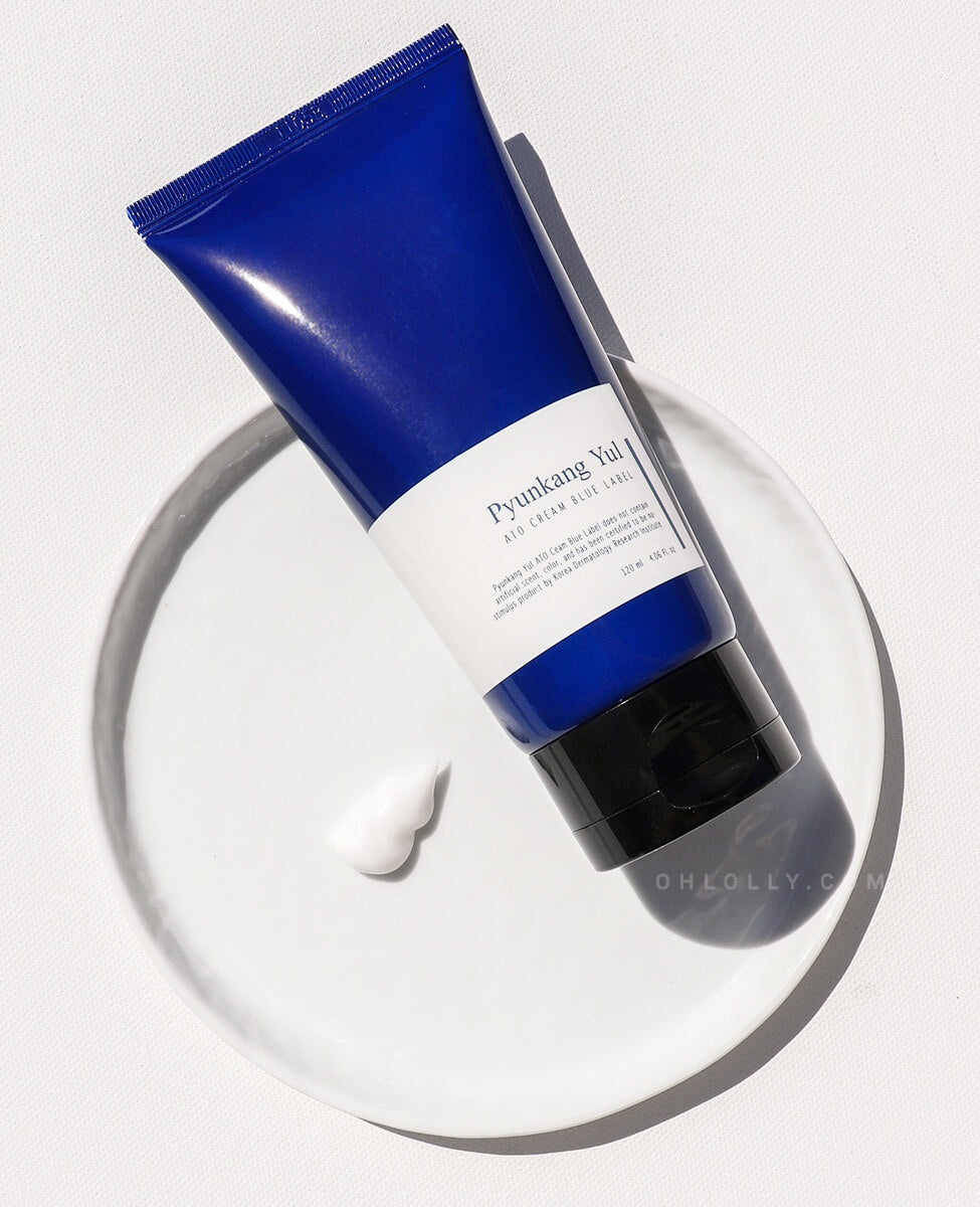 Overhead studio shot of the dark blue Pyunkang Yul ATO Cream Blue Label tube lying on a white porcelain saucer. A perfect dollop of the thick, white cream is visible next to the tube, emphasizing its rich texture and nourishing ceramide and peptide formula.