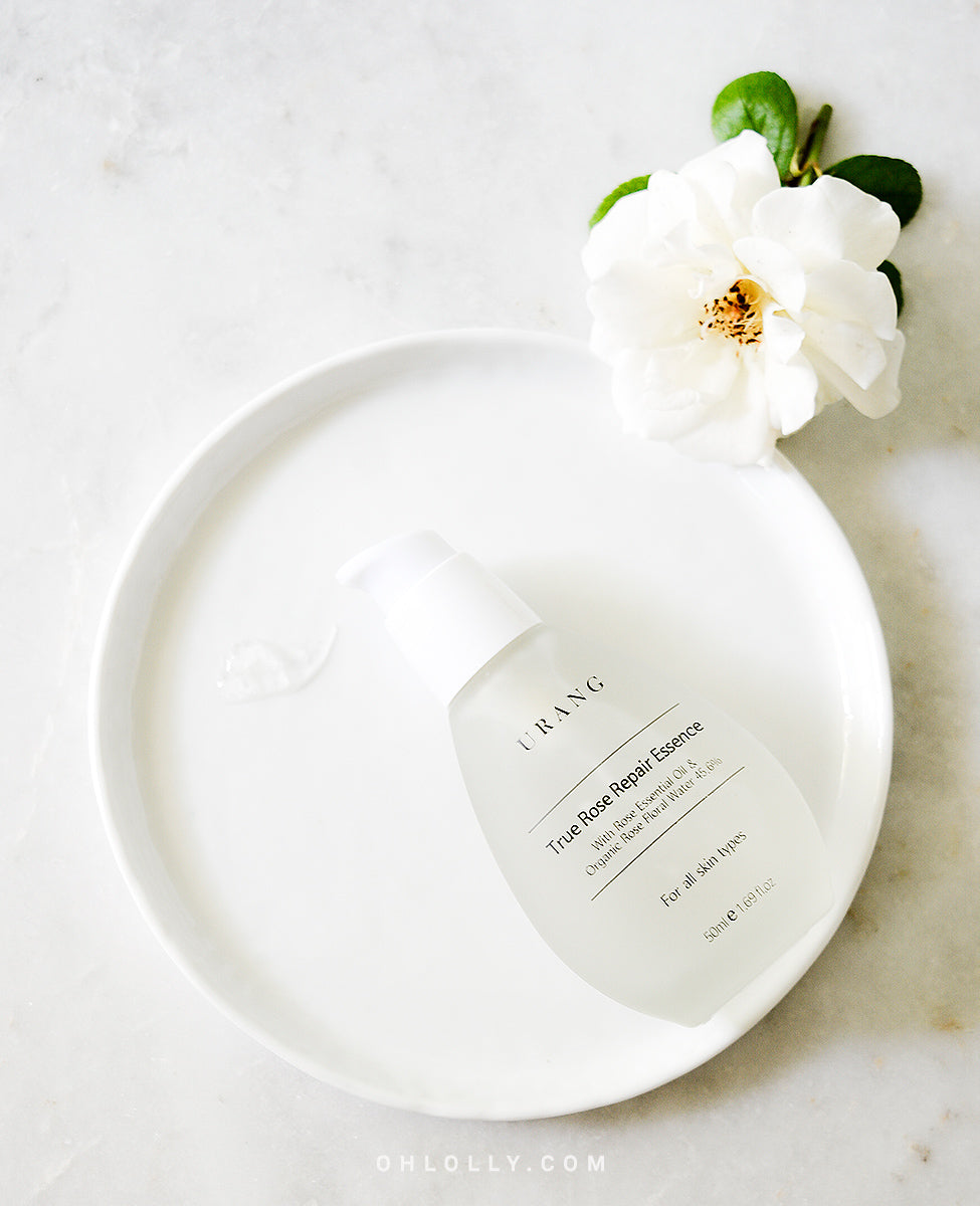 The Urang True Rose Repair Essence pump bottle, resting on a white circular plate with a liquid streak, beside a white rose flower, all on a light marble surface.
