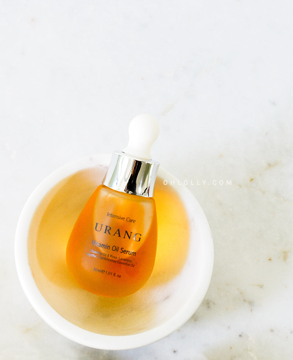 The Urang Vitamin Oil Serum in a frosted glass dropper bottle, filled with bright yellow-orange oil, is displayed upright inside a white ceramic bowl filled with a clear liquid, resting on a white marble surface.
