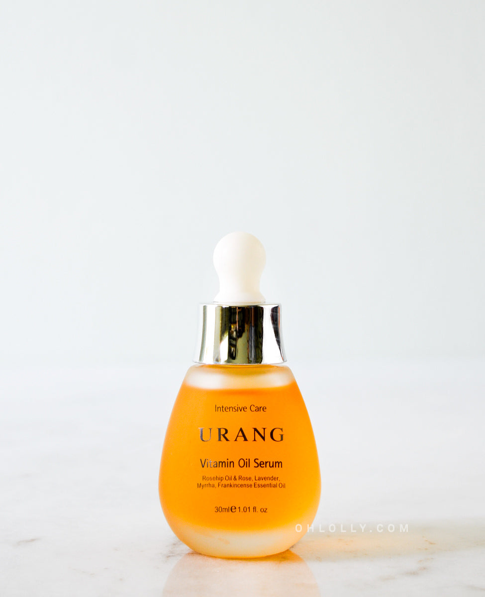The Urang Vitamin Oil Serum in a frosted glass dropper bottle with a white and gold cap, showcasing the yellow-orange oil, sitting on a light marble surface against a white background.
