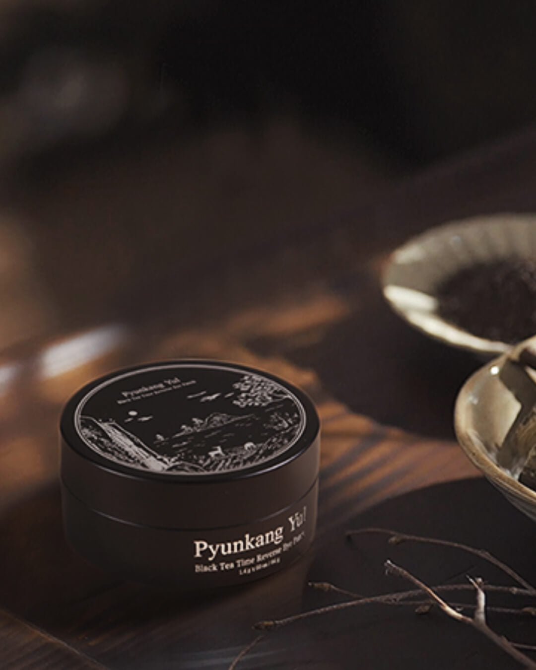 Dark, mood-lit studio shot of the Pyunkang Yul Black Tea Time Reverse Eye Patch jar on a wooden surface. The elegant black jar is in focus, with out-of-focus black tea leaves and bowls in the background, emphasizing the product's luxurious, black tea-infused formula.