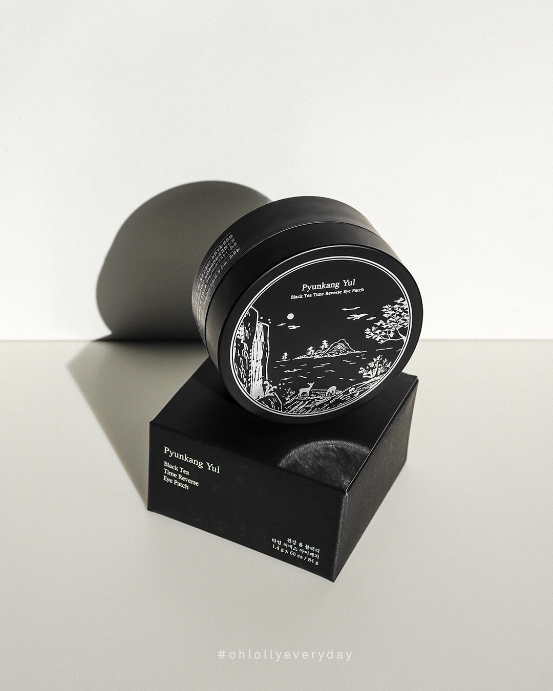 Angled studio shot of the black Pyunkang Yul Black Tea Time Reverse Eye Patch jar sitting on its black, matte outer box. Strong side lighting creates dramatic highlights and shadows, emphasizing the jar's elegant white illustration and the product's premium anti-aging focus.