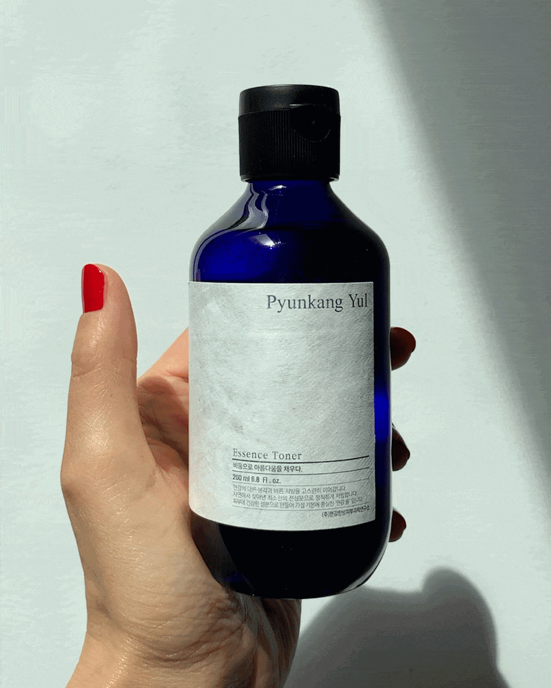 A close-up GIF showing a hand pouring a drop of the Pyunkang Yul Essence Toner from the blue bottle onto the back of another person's hand, highlighting the liquid's clear, slightly viscous texture.