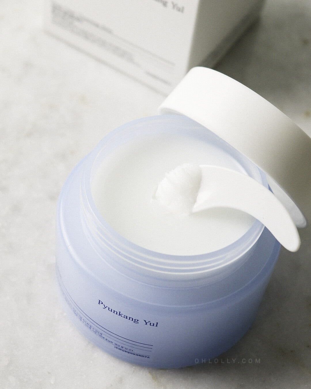 Close-up studio shot of the open Pyunkang Yul Deep Clear Cleansing Balm jar, showing the creamy white balm texture inside. A white spatula rests in the balm, and the jar's lid is angled behind it, emphasizing the product's solid balm-to-oil transformation for deep, soothing cleansing.