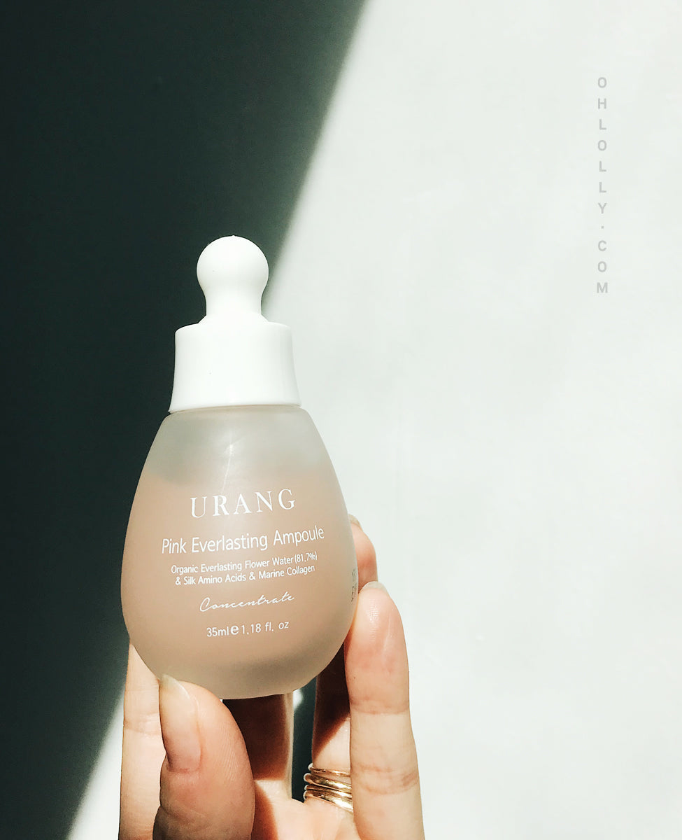 A hand gently holds the frosted pink glass dropper bottle of Urang Pink Everlasting Ampoule, showcasing the minimalist white product label against a bright background with subtle lettering on the right.