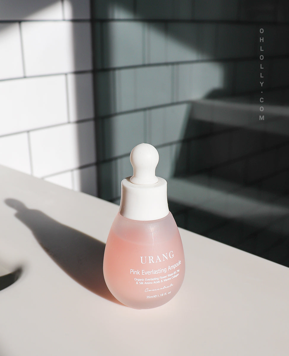 The frosted pink glass bottle of Urang Pink Everlasting Ampoule, highlighted by strong natural sunlight that casts a long shadow on the white surface and textured white background.