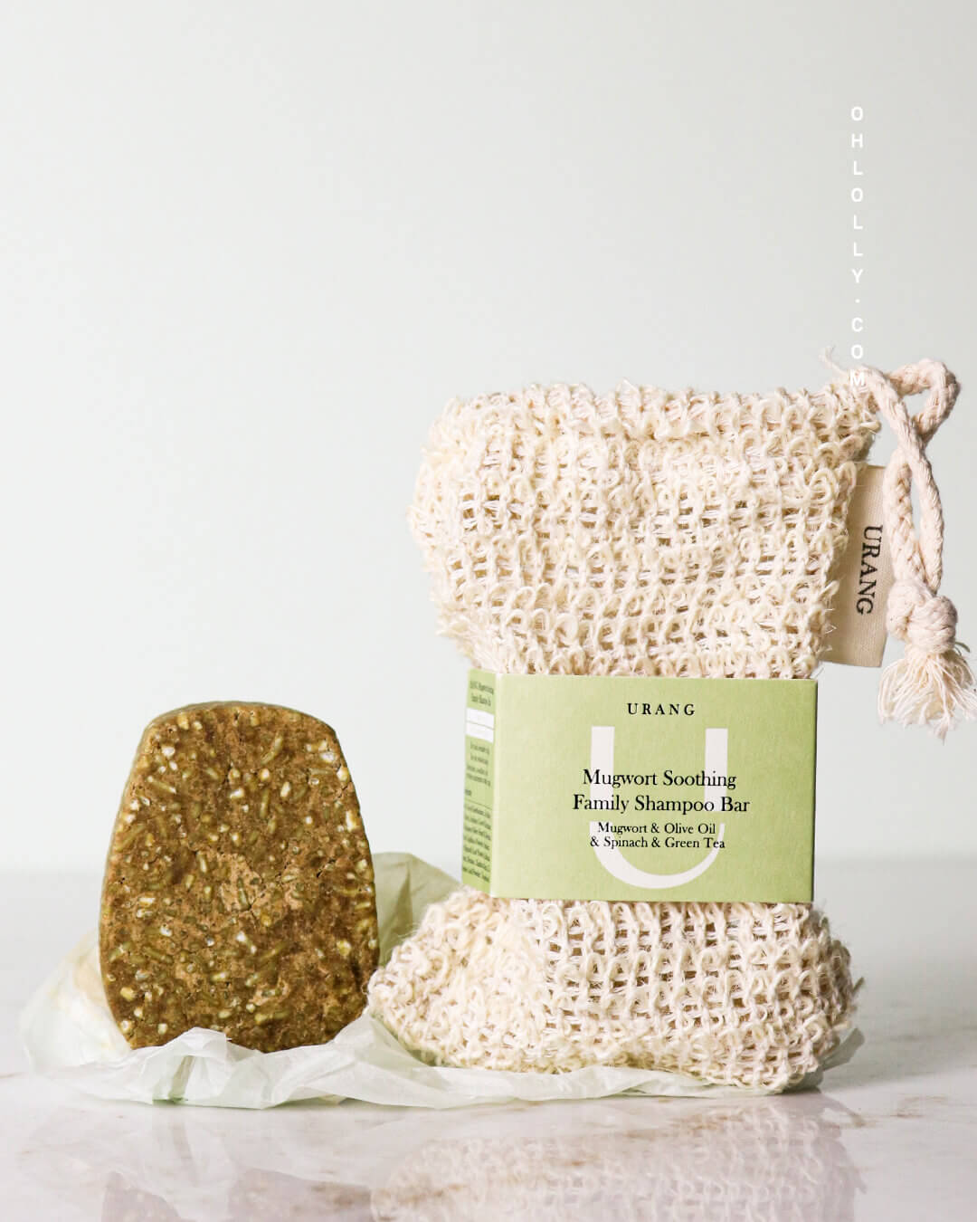 The Urang Mugwort Soothing Family Shampoo Bar, a solid, speckled, green-brown oval, resting next to its woven, light beige cleansing net and its minimalist cardboard packaging sleeve.