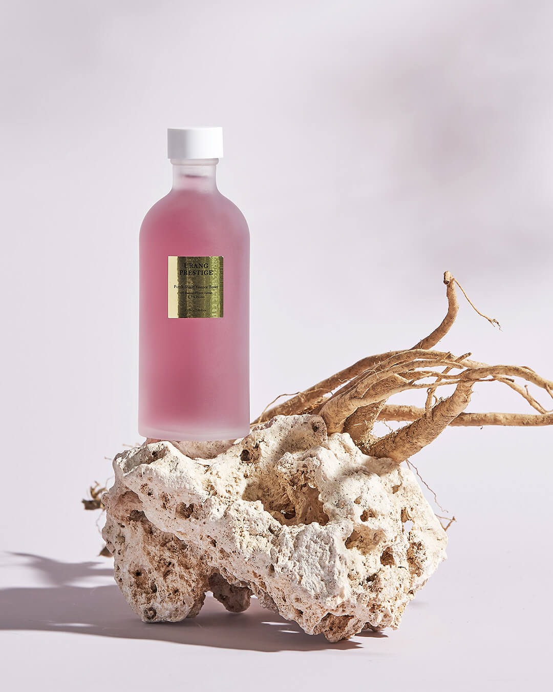 The frosted glass Urang Prestige Purple Shield Essence Toner bottle with purple liquid, standing on a piece of textured white rock, next to dried brown ginseng root.