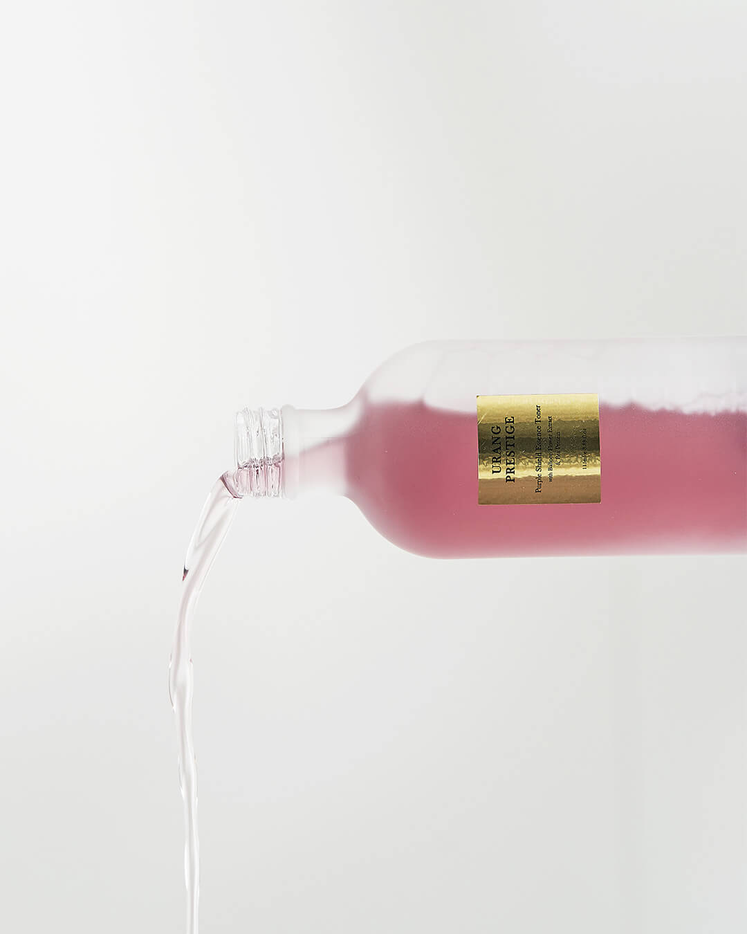 The frosted glass bottle of Urang Prestige Purple Shield Essence Toner is tilted, showing the bright purple, water-like liquid pouring out onto a white background.