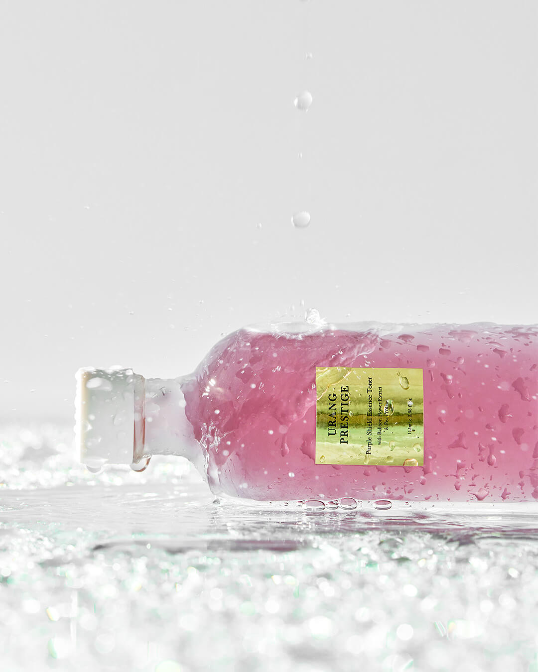 Close-up of the Urang Prestige Purple Shield Essence Toner bottle, lying horizontally and covered in water droplets and splashes, showcasing the bright purple liquid and its golden label.