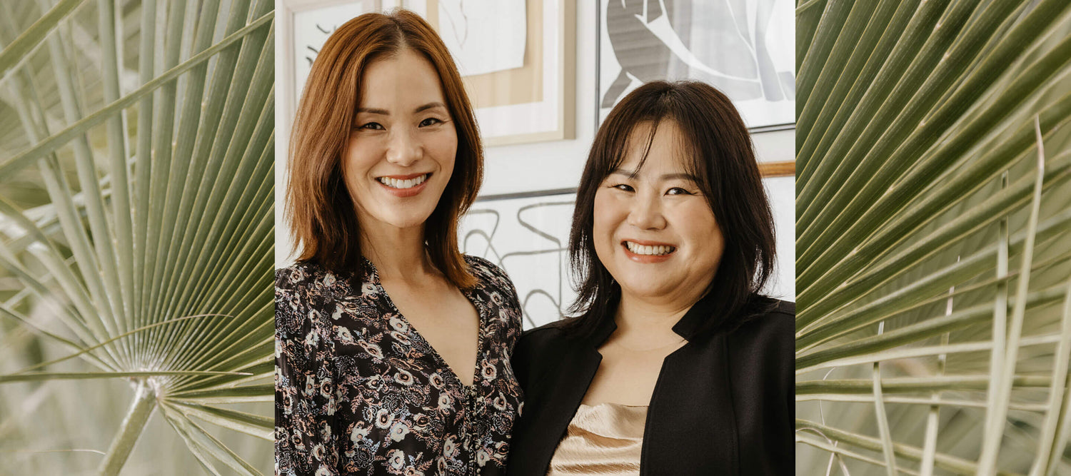Ohlolly founders Sue and Herra smiling, framed by palm leaves. K-beauty experts wishing a Happy Thanksgiving to their community.