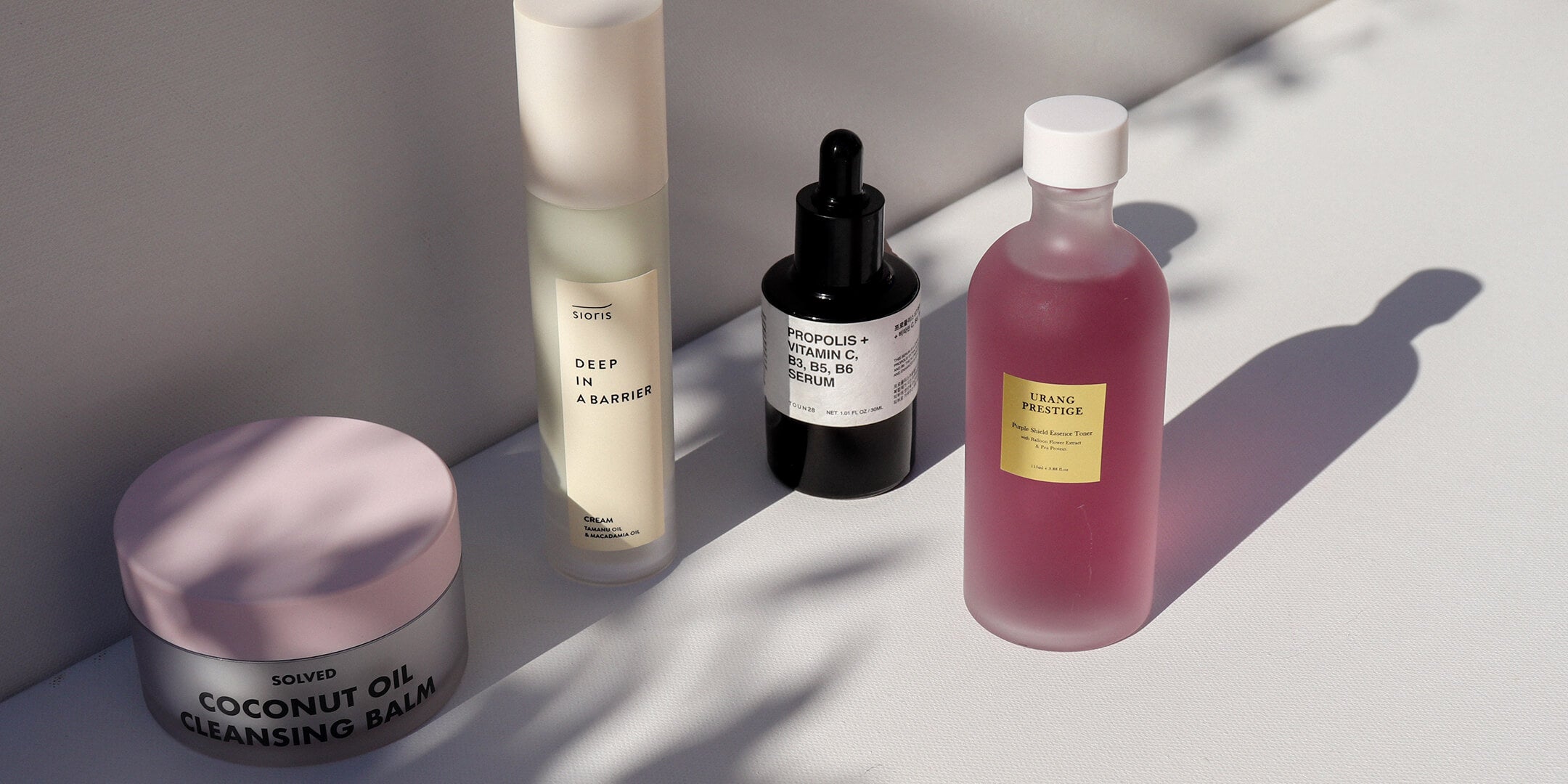 Various skincare products, including a jar of cleansing balm and several serum bottles, arranged on a surface with soft leaf-patterned shadows.