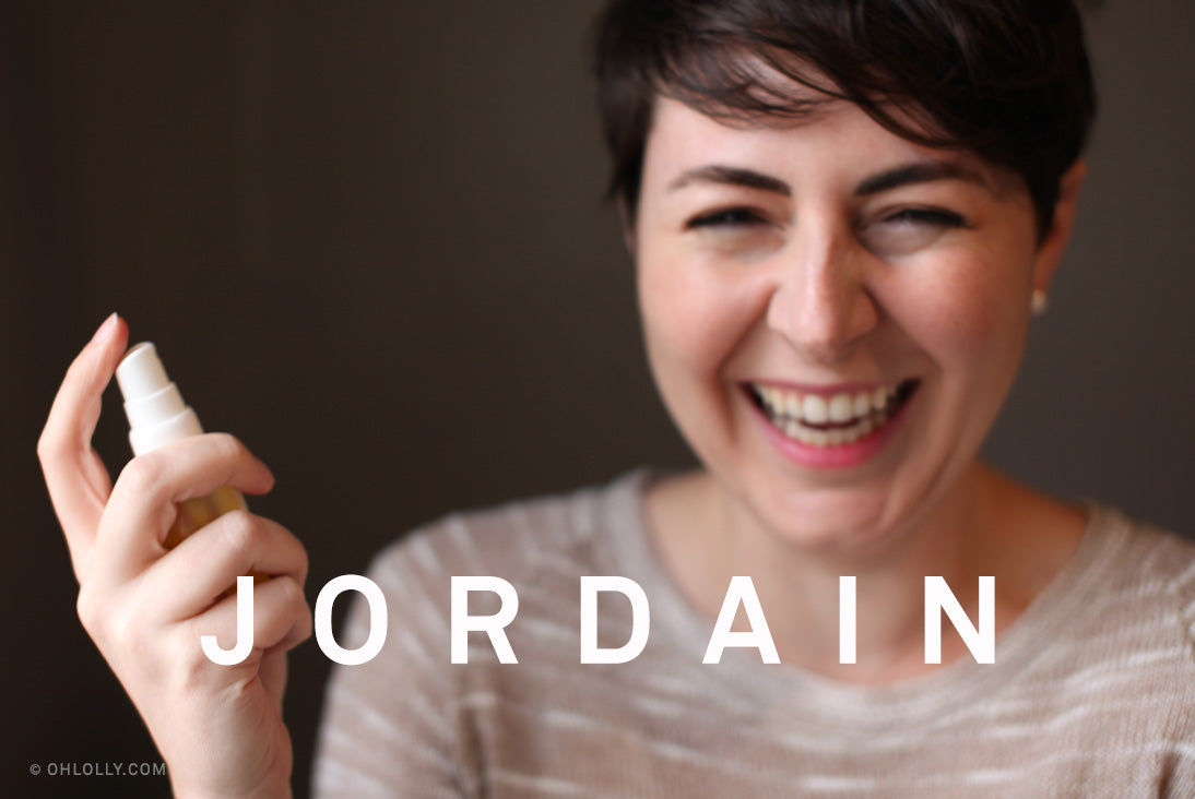 Jordain, a woman with short dark hair, smiling and holding a bottle of spray skincare product near her face.