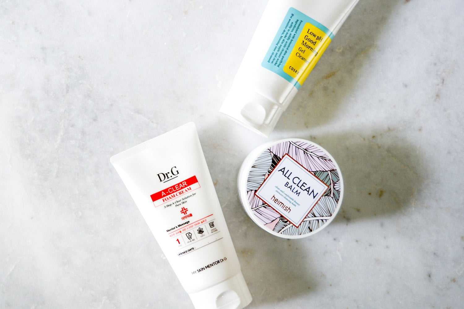 Flat lay photo of three skincare products on a white surface: a white tube with red and black text, a blue and yellow tube, and a round, patterned container.