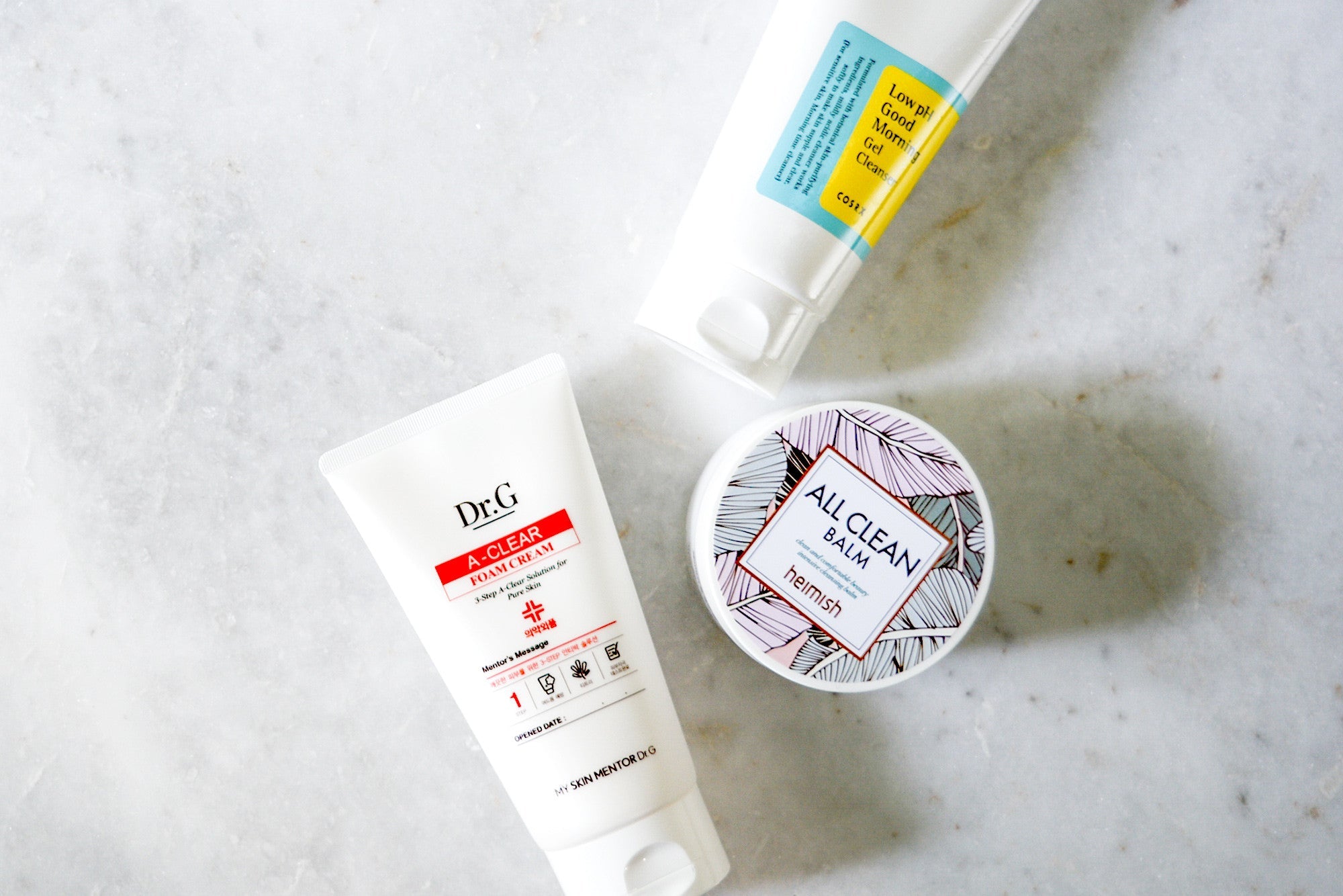 Flat lay photo of three skincare products on a white surface: a white tube with red and black text, a blue and yellow tube, and a round, patterned container.